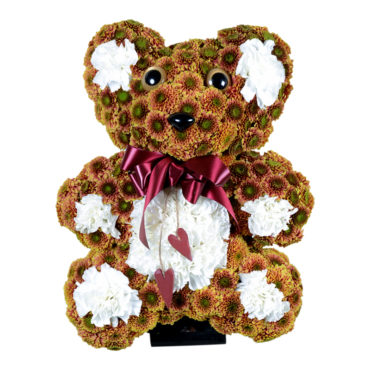 Begravningsblomma i form av en teddy björn gjord av krysantemum. Nallebjörnen har en rosett runt halsen och är brun med vita detaljer. Dekorationen är i mitten av en vit bakgrund och är ett personligt arrangemang.