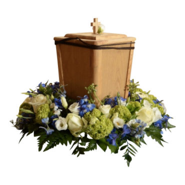 Brun trä urn med kristet kors på ovansidan. Urnen är i mitten av en urndekoration med flertal begravningsblommor som formar en urnkrans i färgerna svalkande blå,vit och svaga gröna toner.